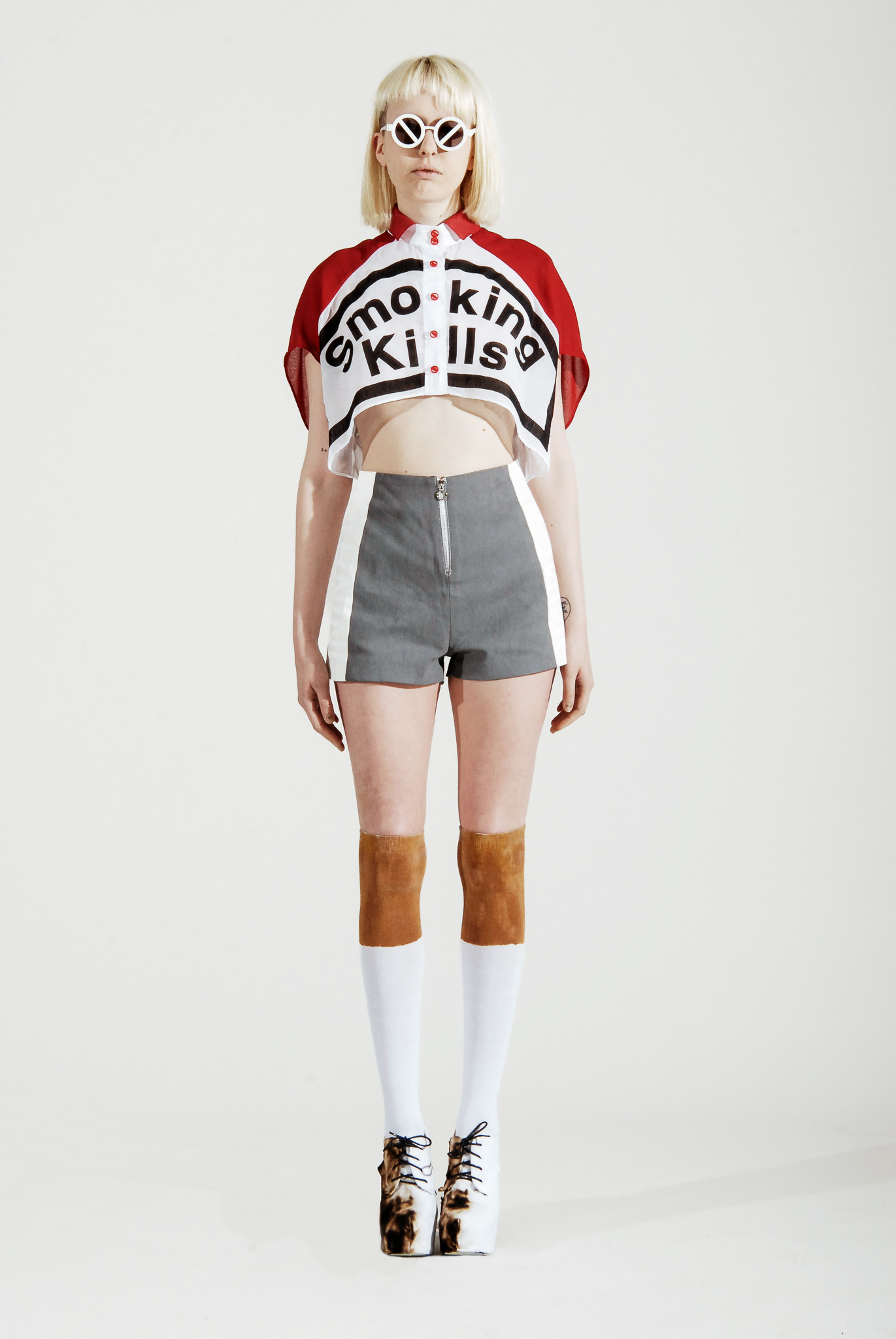 Front view of Look 01 from Smoking Kills, featuring a red and white cropped shirt, gray shorts, knee-high socks, and white platform shoes.